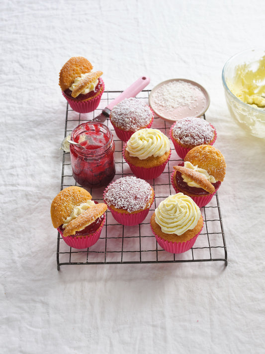 Fairy cakes on a wire rack with a pot of jam, a pink spoon, a plate of icing sugar and a bowl of buttercream frosting off to the side